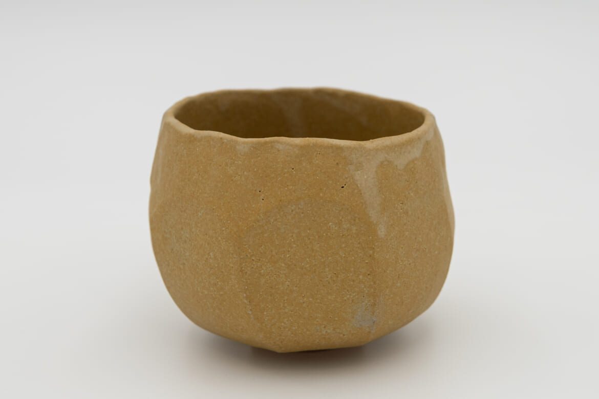 Holding, a hand-formed ochre stoneware chawan with a compact rounded silhouette and softly undulating rim, by Celeste Ortelee.
