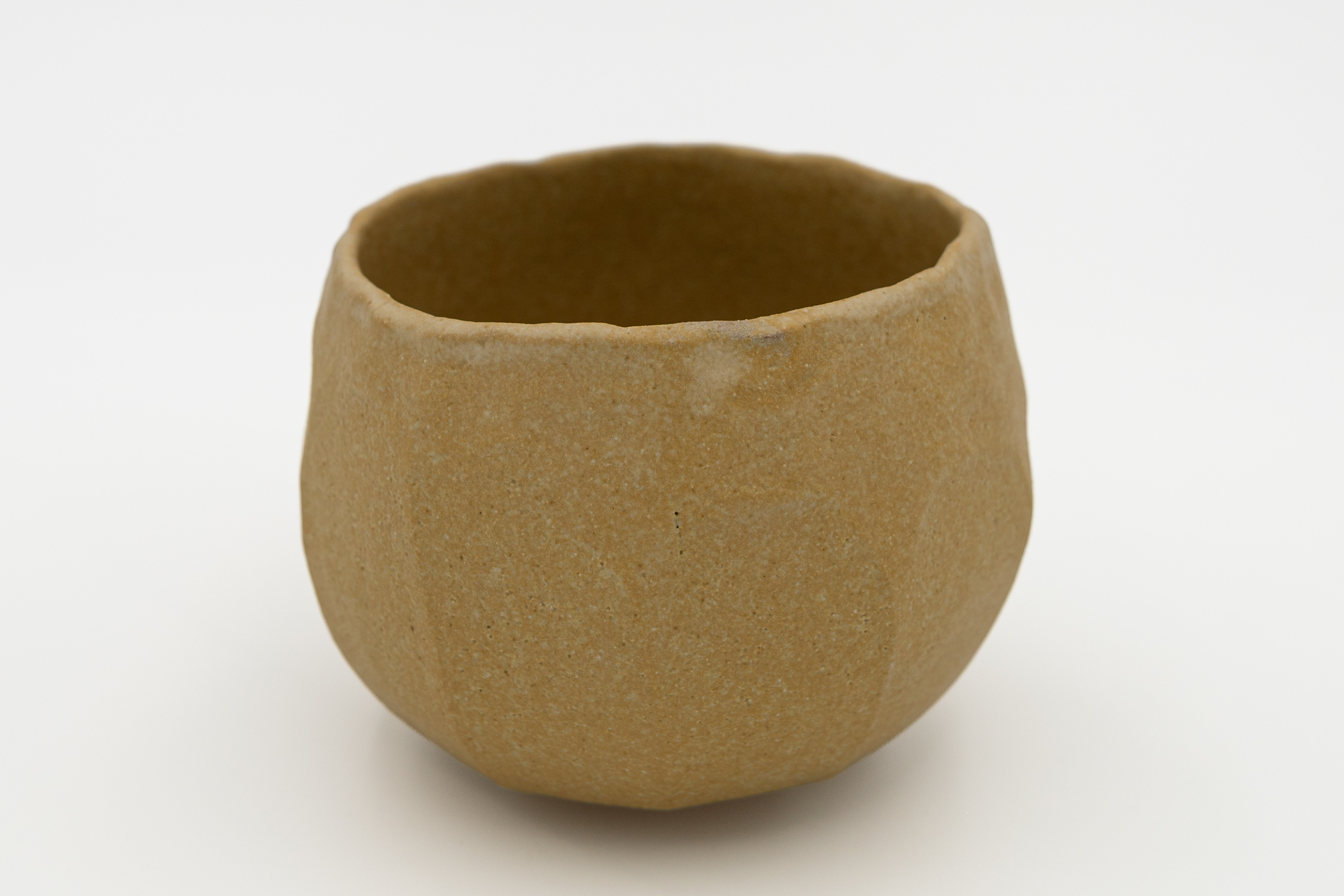 Holding, an ochre-glazed ceramic tea bowl seen from a second angle, showing its softly irregular rim and hand-shaped surface.
