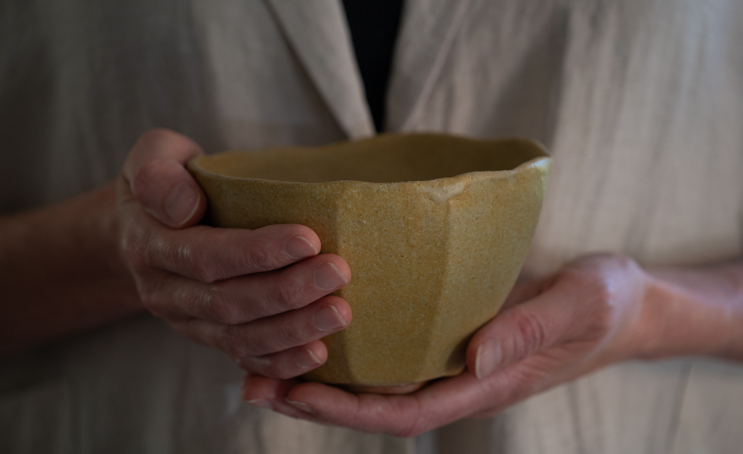 Hands holding Unfolding, an ochre-glazed stoneware chawan by Celeste Ortelee, with one hand supporting the foot.